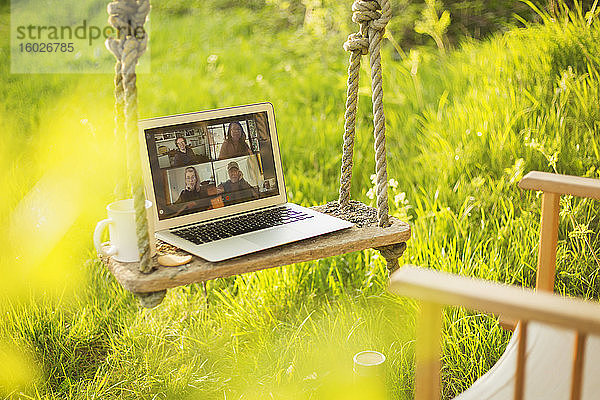 Video-Chat mit Kollegen am Laptop-Bildschirm auf einer rustikalen Schaukel im Garten