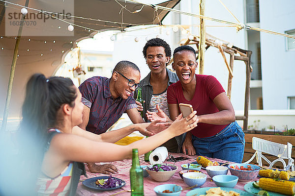 Glückliche junge Freunde  die ein Smartphone benutzen und am Balkontisch essen