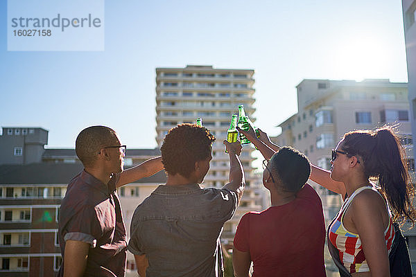 Junge Freunde stoßen auf Bierflaschen auf dem sonnigen städtischen Dachbalkon an