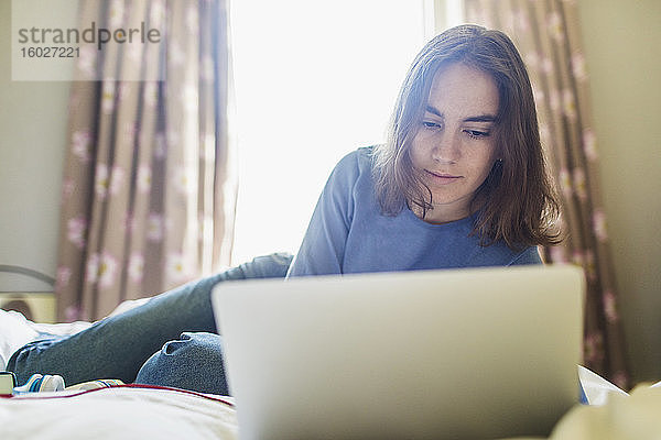 Teenager-Mädchen benutzt Laptop auf dem Bett im sonnigen Schlafzimmer