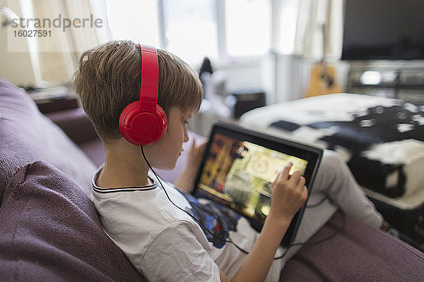 Junge mit Kopfhörer und digitalem Tablet spielt Videospiel auf dem Sofa