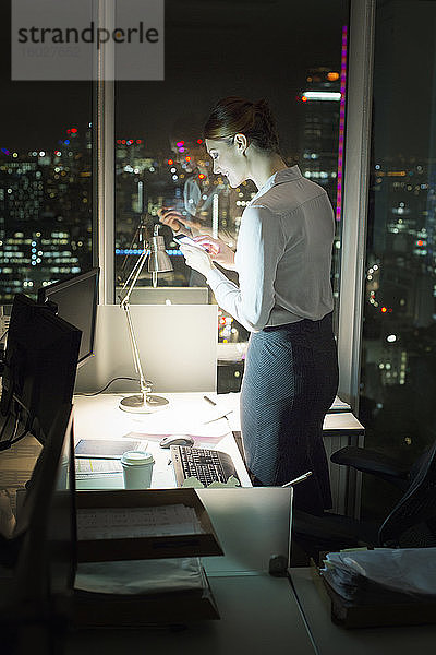 Geschäftsfrau  die nachts im Büro Textnachrichten mit dem Handy verschickt