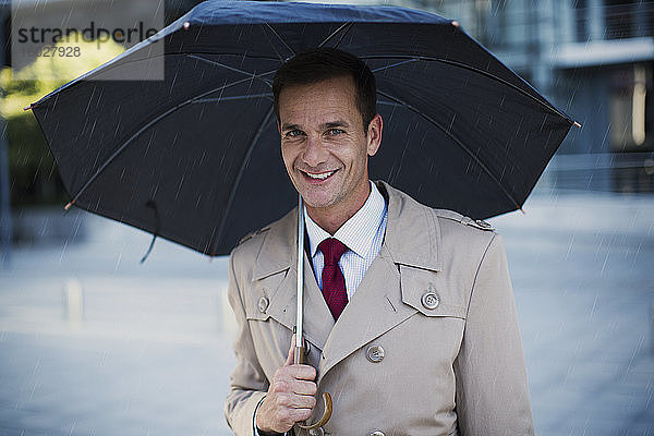 Lächelnder Geschäftsmann im Trenchcoat unterm Regenschirm