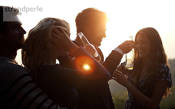 Zwei Männer und zwei Frauen stehen bei Sonnenuntergang im Freien  halten Bierflaschen in der Hand und lächeln.