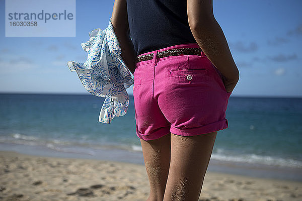 Rückansicht einer Frau in rosa Hotpants  die an einem Sandstrand am Meer steht.