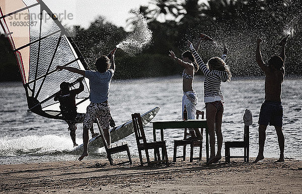 Kleine Gruppe von Männern und Frauen  die auf einem Sandstrand um Tisch und Stühle herum stehen  die Arme erhoben  Flaschen haltend  einen Windsurfer beobachtend.