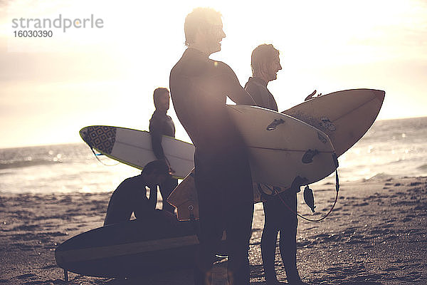 Vier Männer in Neoprenanzügen stehen an einem Sandstrand und tragen Surfbretter.