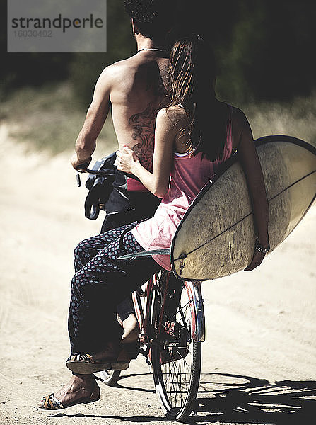 Rückansicht von Mann und Frau auf einem Fahrrad  Frau sitzt auf einem Gepäckträger und hält ein Surfbrett.