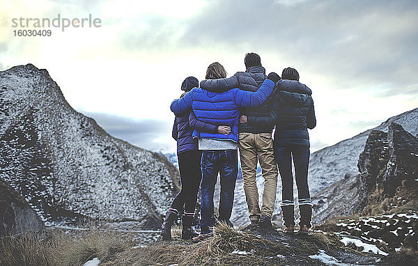 Rückansicht von vier Menschen  die Arm in Arm auf einem Berg stehen  schneebedeckte Gipfel in der Ferne.