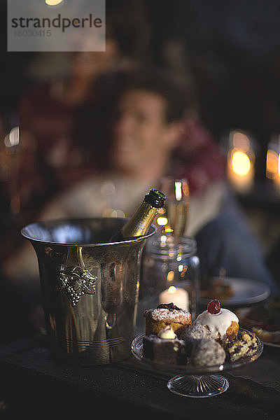 Nahaufnahme der Champagnerflasche im Weinkühler aus Metall  Kuchenstand aus Glas mit Kuchenauswahl  Person im Hintergrund.