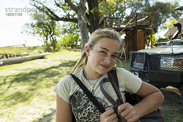 Ein zwölfjähriges Mädchen mit Taschen in einem Wildreservatscamp  im Hintergrund Gebäude und Boote.