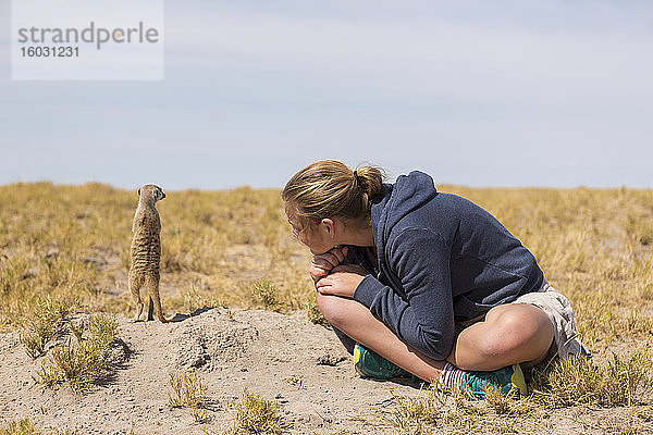 12-jähriges Mädchen sitzt und beobachtet  wie Erdmännchen aus ihren Höhlen in der Kalahari-Wüste auftauchen.