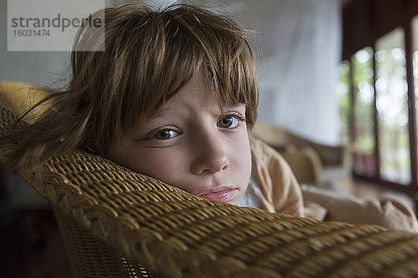 Ein müder fünfjähriger Junge  der sich in einem Hotel in einem Korbstuhl ausruht und in die Kamera schaut.