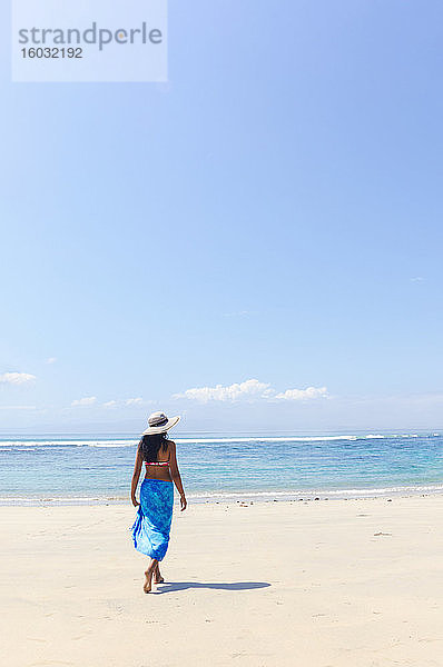 Eine junge Frau mit Sonnenhut und Schal  die an einem unberührten tropischen Strand auf der Insel Bali spazieren geht  Indonesien  Südostasien  Asien