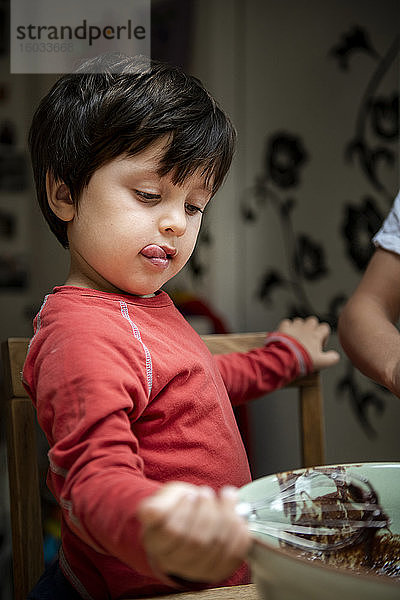 Ein Junge mit schwarzen Haaren sitzt an einem Küchentisch und backt Schokoladenkuchen.