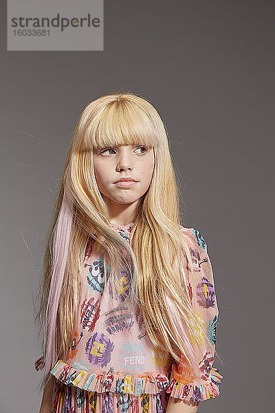 Porträt eines Mädchens mit langen blonden Haaren  das ein rosafarbenes Rüschen-Pom-Pom-Monsterkleid trägt und in die Kamera schaut  auf grauem Hintergrund.