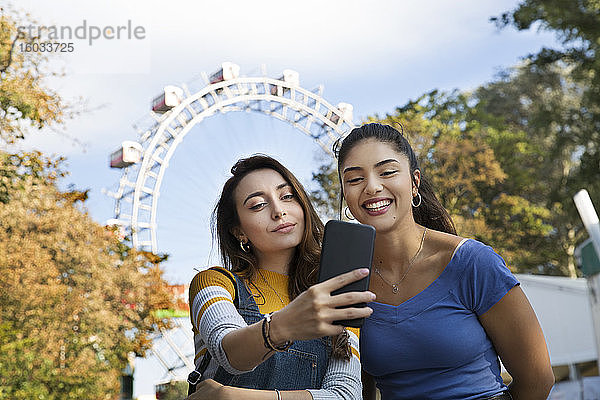 Zwei junge Frauen mit langen braunen Haaren  die in einem Park in der Nähe eines Riesenrads stehen und sich mit einem Mobiltelefon selbstständig machen.