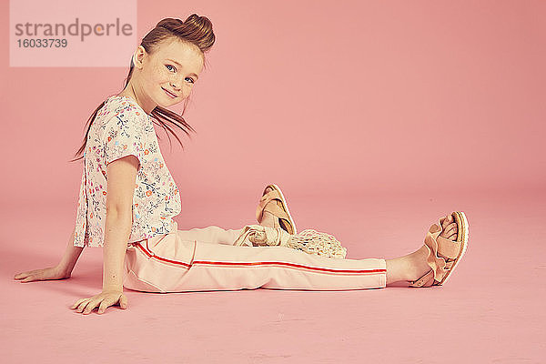 Porträt eines brünetten Mädchens auf rosa Hintergrund  mit blumigem Oberteil und zartrosa Hose  das auf dem Boden sitzt und in die Kamera schaut.
