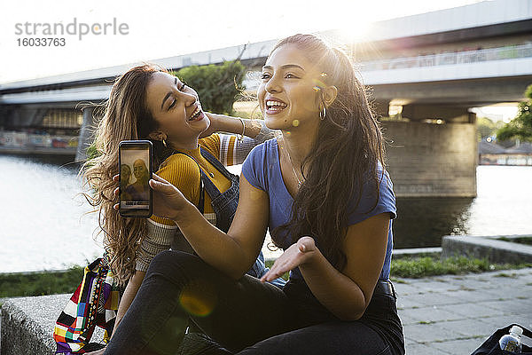 Zwei junge Frauen mit langen braunen Haaren sitzen am Flussufer  halten ein Handy in der Hand und lächeln.