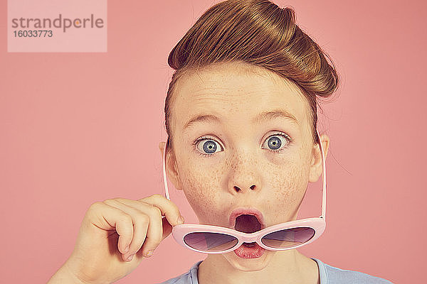 Porträt eines brünetten Mädchens auf rosa Hintergrund  das mit offenem Mund über die Sonnenbrille in die Kamera schaut.