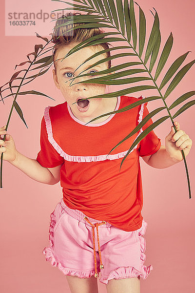 Porträt eines brünetten Mädchens auf rotem T-Shirt  versteckt hinter Palmblatt  auf rosa Hintergrund  in die Kamera blickend.