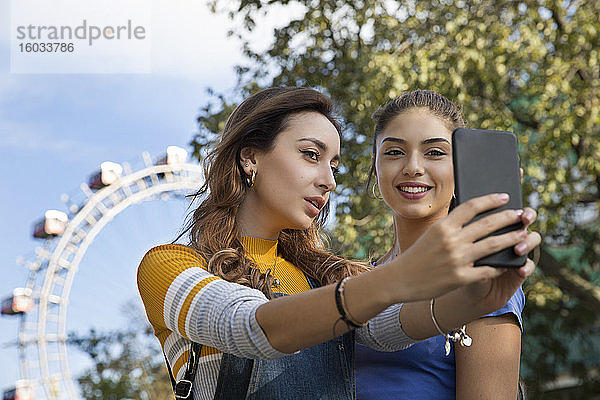 Zwei junge Frauen mit langen braunen Haaren  die in einem Park in der Nähe eines Riesenrads stehen und sich mit einem Mobiltelefon selbstständig machen.