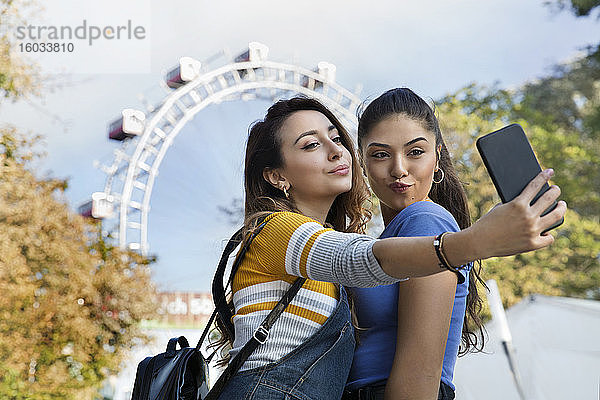 Zwei junge Frauen mit langen braunen Haaren  die in einem Park in der Nähe eines Riesenrads stehen und sich mit einem Mobiltelefon selbstständig machen.