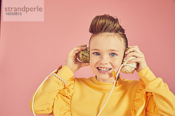 Porträt eines brünetten Mädchens in gelbem Top  das ein Muscheltelefon hält  auf rosa Hintergrund  in die Kamera blickend.