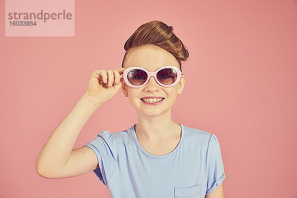 Porträt eines brünetten Mädchens mit blauem T-Shirt und Sonnenbrille auf rosa Hintergrund  das in die Kamera schaut.