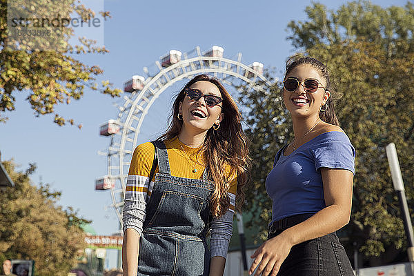 Zwei junge Frauen mit langen braunen Haaren stehen in einem Park in der Nähe eines Riesenrads  tragen eine Sonnenbrille und lächeln in die Kamera.
