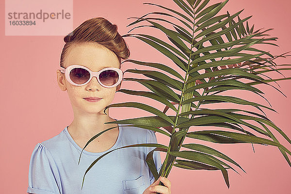 Porträt eines brünetten Mädchens mit blauem T-Shirt und Sonnenbrille auf rosa Hintergrund  das ein Palmblatt hält und in die Kamera schaut.