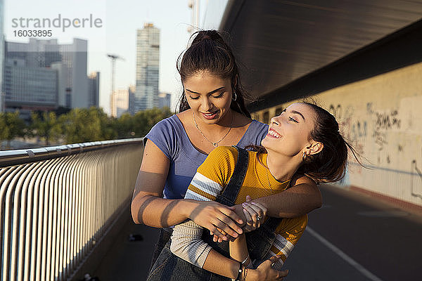 Zwei junge Frauen mit langen braunen Haaren stehen auf einer Stadtbrücke  umarmen sich und lächeln.
