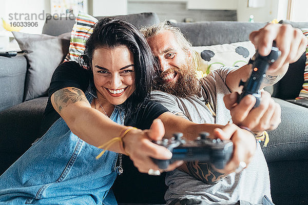 Bärtiger tätowierter Mann mit langen brünetten Haaren und Frau mit langen braunen Haaren sitzen auf einem Sofa und lächeln beim Konsolenspiel.