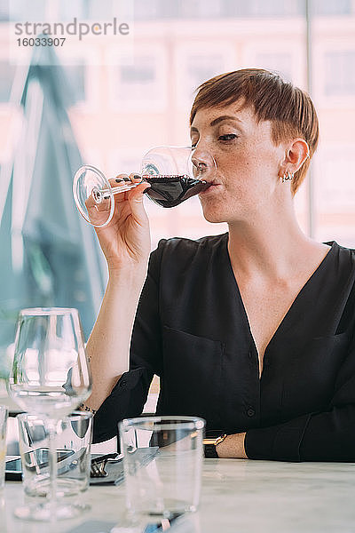 Junge Frau mit kurzen Haaren in schwarzem Top  die in einer Bar am Tisch sitzt und Rotwein trinkt.