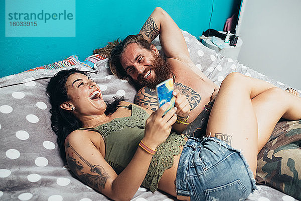 Bärtiger tätowierter Mann mit langen brünetten Haaren und Frau mit langen braunen Haaren liegen lachend auf einem Bett.