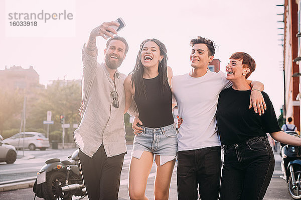 Zwei junge Frauen und Männer in legerer Kleidung auf einem Dach stehend  die sich mit einem Mobiltelefon selbstständig machen.