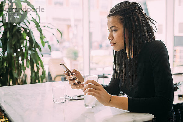 Junge Frau mit Dreadlocks  in schwarzem T-Shirt  sitzt am Tisch in einer Bar und benutzt ein Mobiltelefon.