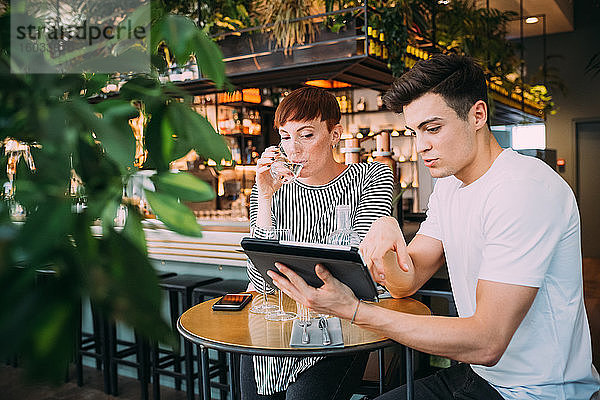 Junge Frau und Mann sitzen an einem Tisch in einer Bar und schauen auf ein digitales Tablett.