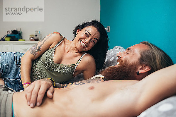 Bärtiger tätowierter Mann mit langen brünetten Haaren und Frau mit langen braunen Haaren liegen auf einem Bett und lächeln sich an.
