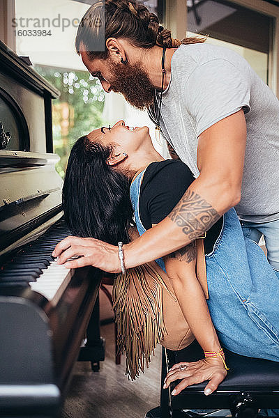 Bärtiger tätowierter Mann mit langen brünetten Haaren  der sich über eine lächelnde Frau beugt  die Klavier spielt.