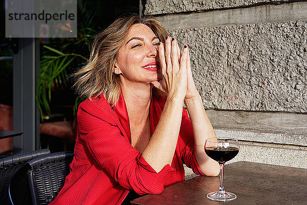Porträt einer sitzenden Frau mit langen brünetten Haaren  mit roter Jacke  die Hände gefaltet  ein Glas Wein auf einem Tisch