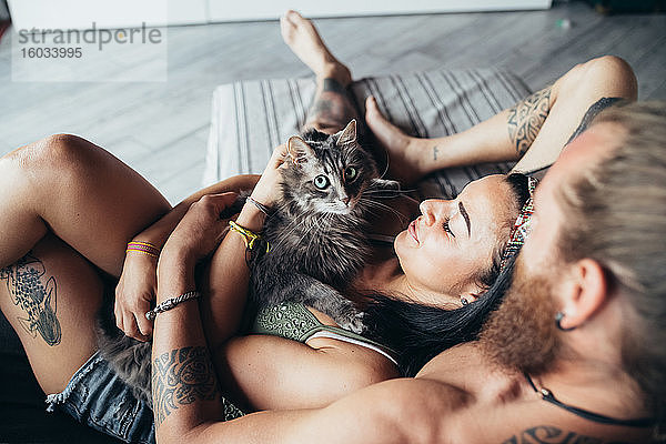 Bärtig tätowierter Mann mit langen brünetten Haaren und Frau mit langen braunen Haaren  die mit einer flauschigen grauen Katze auf einem Sofa kuschelt.
