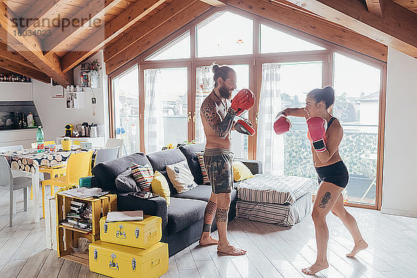 Bärtig tätowierter Mann mit langen brünetten Haaren und Frau mit langen braunen Haaren stehen drinnen und üben Kickboxen.