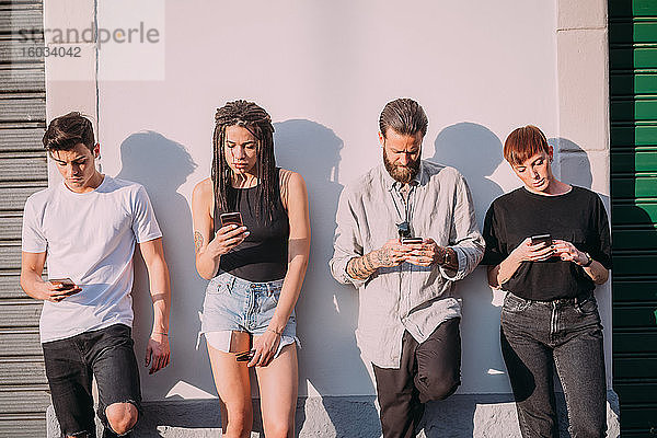 Zwei junge Frauen und Männer in legerer Kleidung  die an der Wand lehnen und Mobiltelefone benutzen.