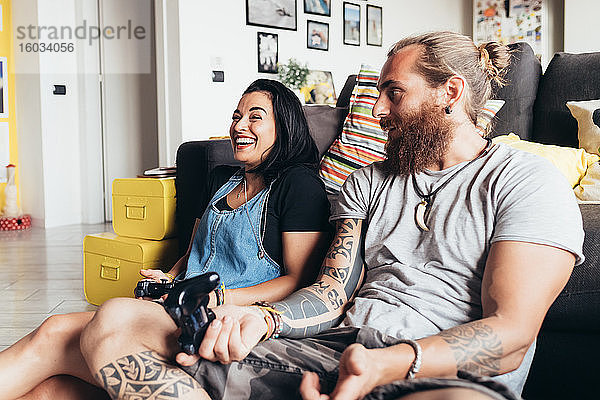 Bärtiger tätowierter Mann mit langen brünetten Haaren und Frau mit langen braunen Haaren sitzen auf einem Sofa und lächeln beim Konsolenspiel.