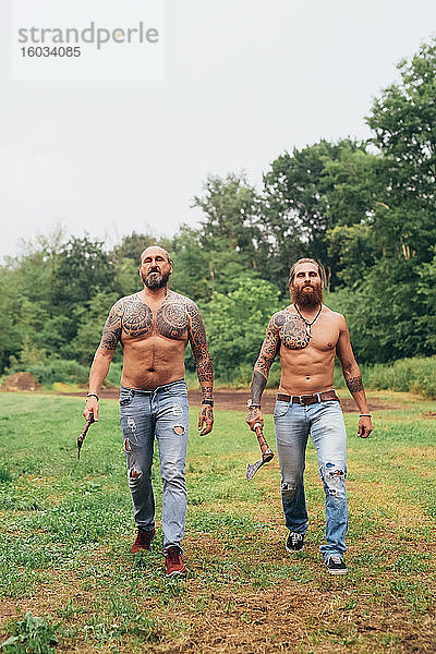 Zwei bärtige tätowierte Männer mit langen brünetten Haaren  ohne Hemd und in Jeans  die über eine Wiese gehen und Äxte tragen.