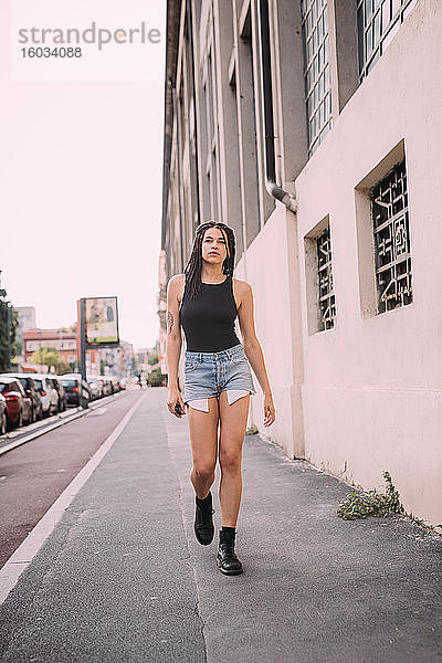 Porträt einer jungen Frau mit dunkelbraunem Haar  schwarzer Weste und Jeans-Shorts  die die Straße entlang geht.