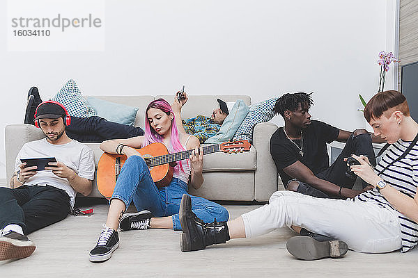 Gruppe junger Männer und Frauen  die auf dem Boden des Wohnzimmers sitzen und ihre Mobiltelefone überprüfen  Frau mit langen rosa Haaren  die Gitarre spielt.