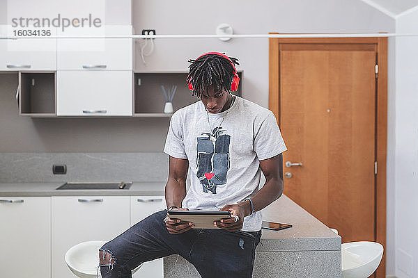Junger Mann mit kurzen Dreadlocks sitzt in einer Küche  trägt Kopfhörer und hält ein digitales Tablett in der Hand.