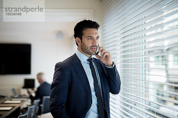 Bärtiger Geschäftsmann in Anzug und Krawatte  der am Fenster steht und ein Mobiltelefon benutzt.